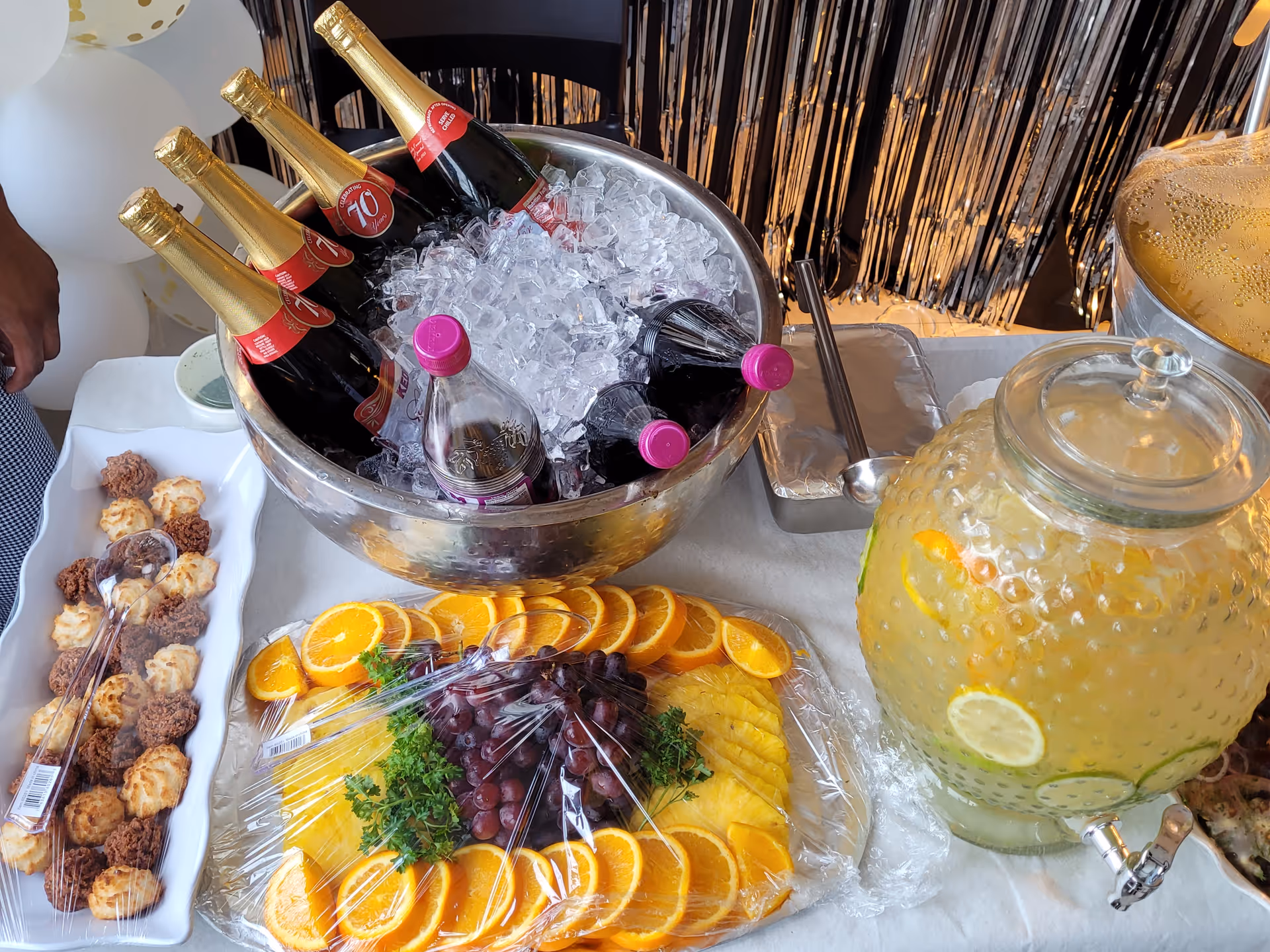 Table with a beverage and snack buffet featuring a bowl of champagne bottles on ice, a large glass dispenser of citrus drink, a platter of sliced fruit and trays of small pastries.