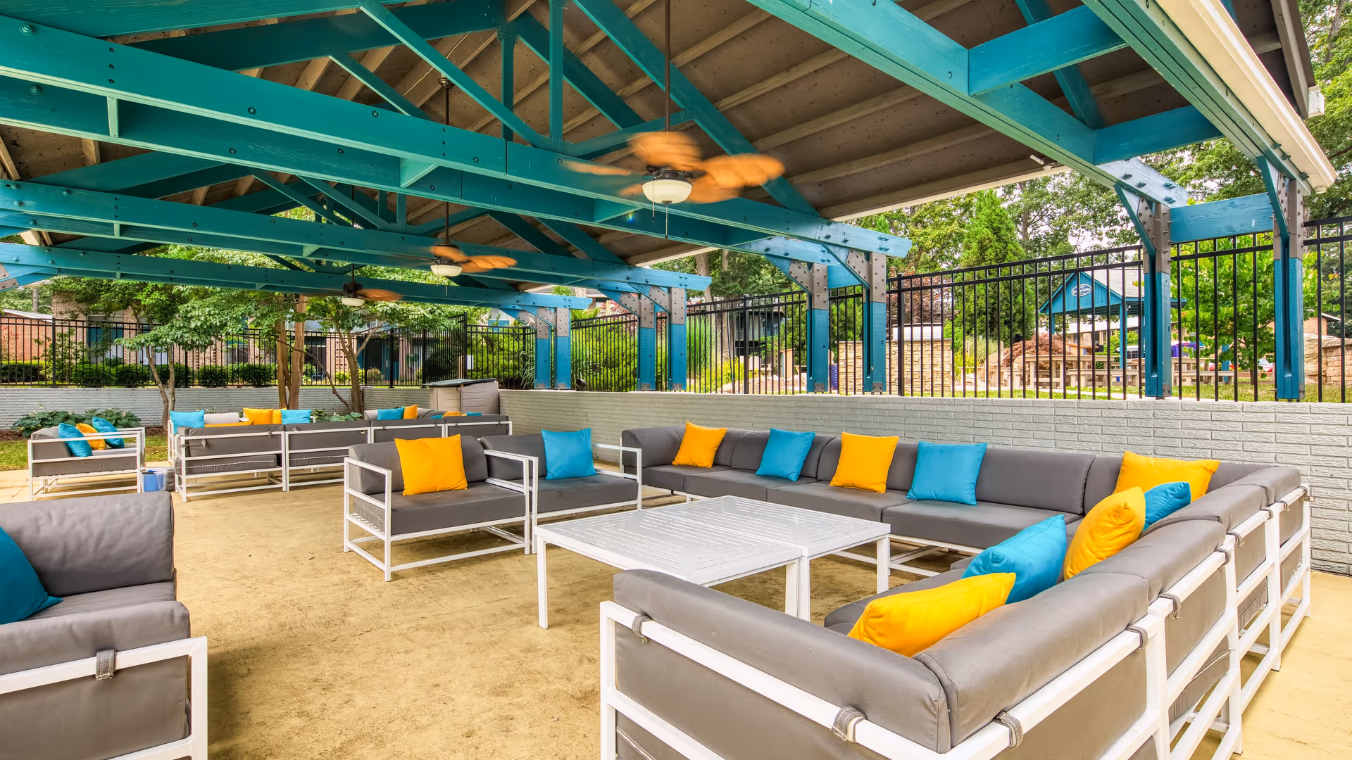 Outdoor covered seating area with gray cushioned sofas and chairs accented with blue and yellow pillows, under a teal wooden roof structure with ceiling fans, surrounded by a black metal fence and greenery.