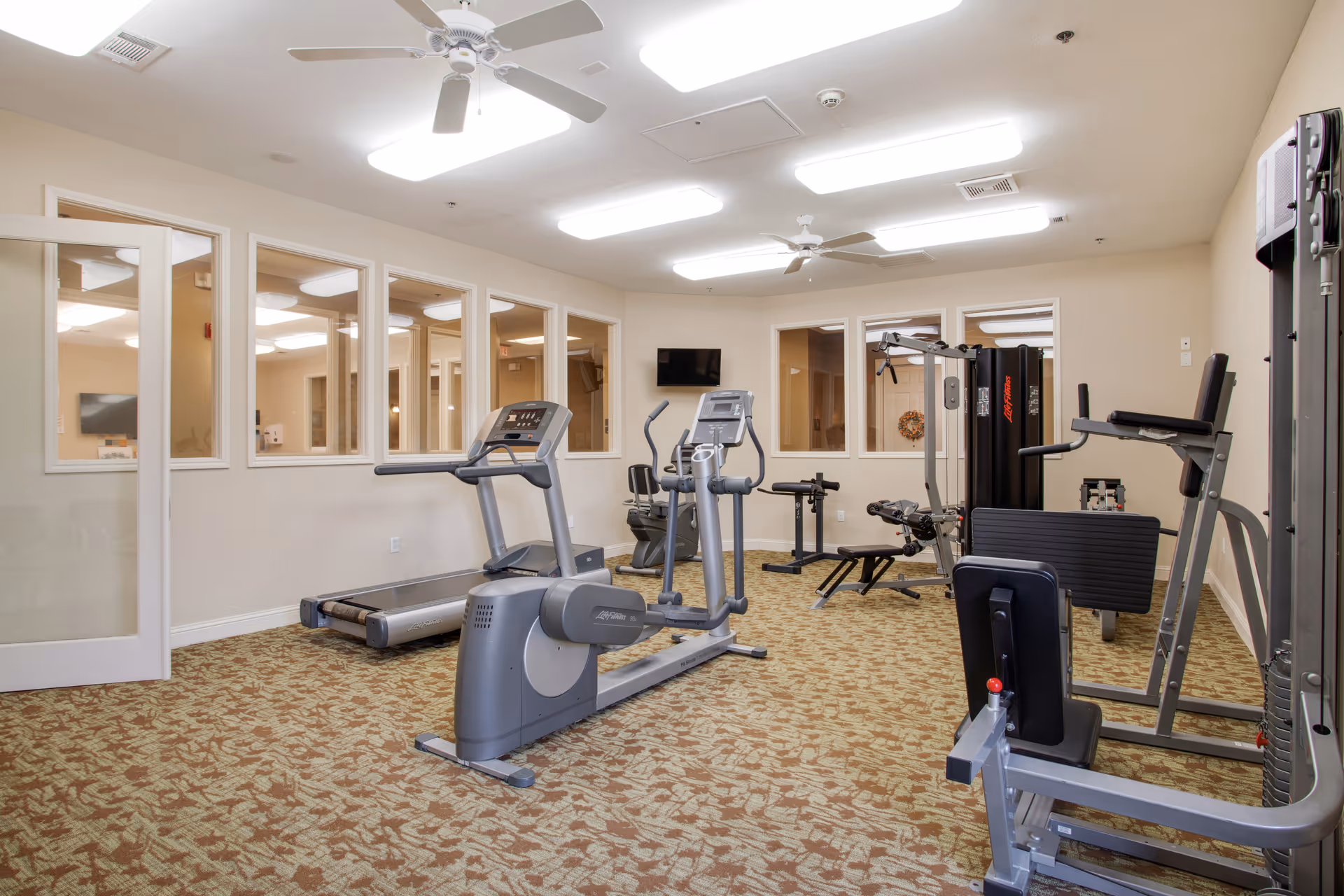Interior view of a fitness room in a senior living facility with exercise equipment including a treadmill, elliptical machine, weight machines, and a stationary bike. The room has beige walls, patterned carpet, ceiling fans, and multiple windows looking into adjacent rooms.