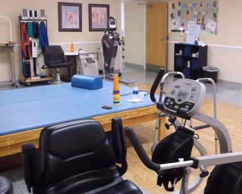 Therapy gym with exercise bike, blue mat, and medical equipment in a senior facility.