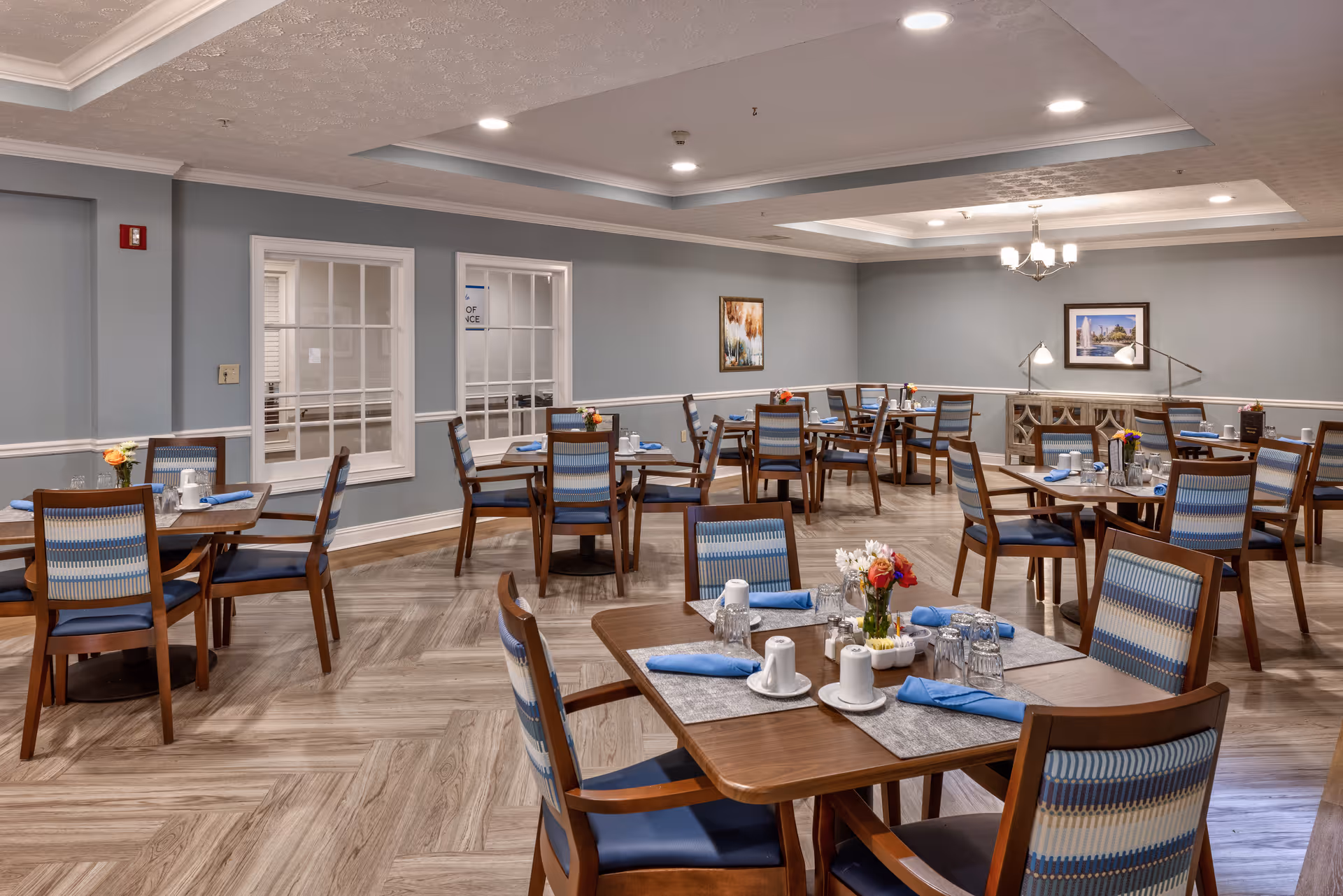 A spacious dining room with multiple wooden tables and chairs featuring blue and beige striped upholstery. Each table is set with blue napkins, glasses, and small flower arrangements. The room has light gray walls, wood-patterned flooring, recessed lighting, and framed artwork on the walls.