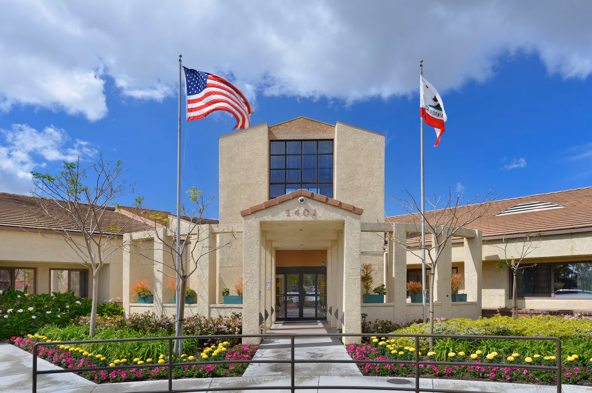 Front exterior view of Walnut Village Orange County Retirement Community building with two flagpoles displaying the American flag and the California state flag, surrounded by landscaped flower beds and a clear blue sky with some clouds.