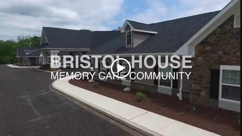 Exterior view of a single-story building with stone and siding walls, multiple windows, and a dark shingled roof. A paved driveway and a concrete sidewalk run alongside the building, with small landscaped areas containing plants and mulch. The sky is partly cloudy.