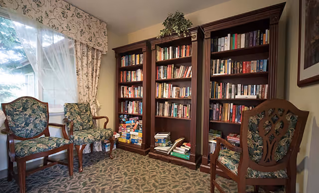 A cozy reading room with three wooden chairs upholstered in floral fabric, positioned near a large window with sheer curtains and floral valance. Against the wall are three tall wooden bookshelves filled with books and board games, with a small plant on top of the middle shelf. The room has patterned carpet flooring and soft yellow walls.