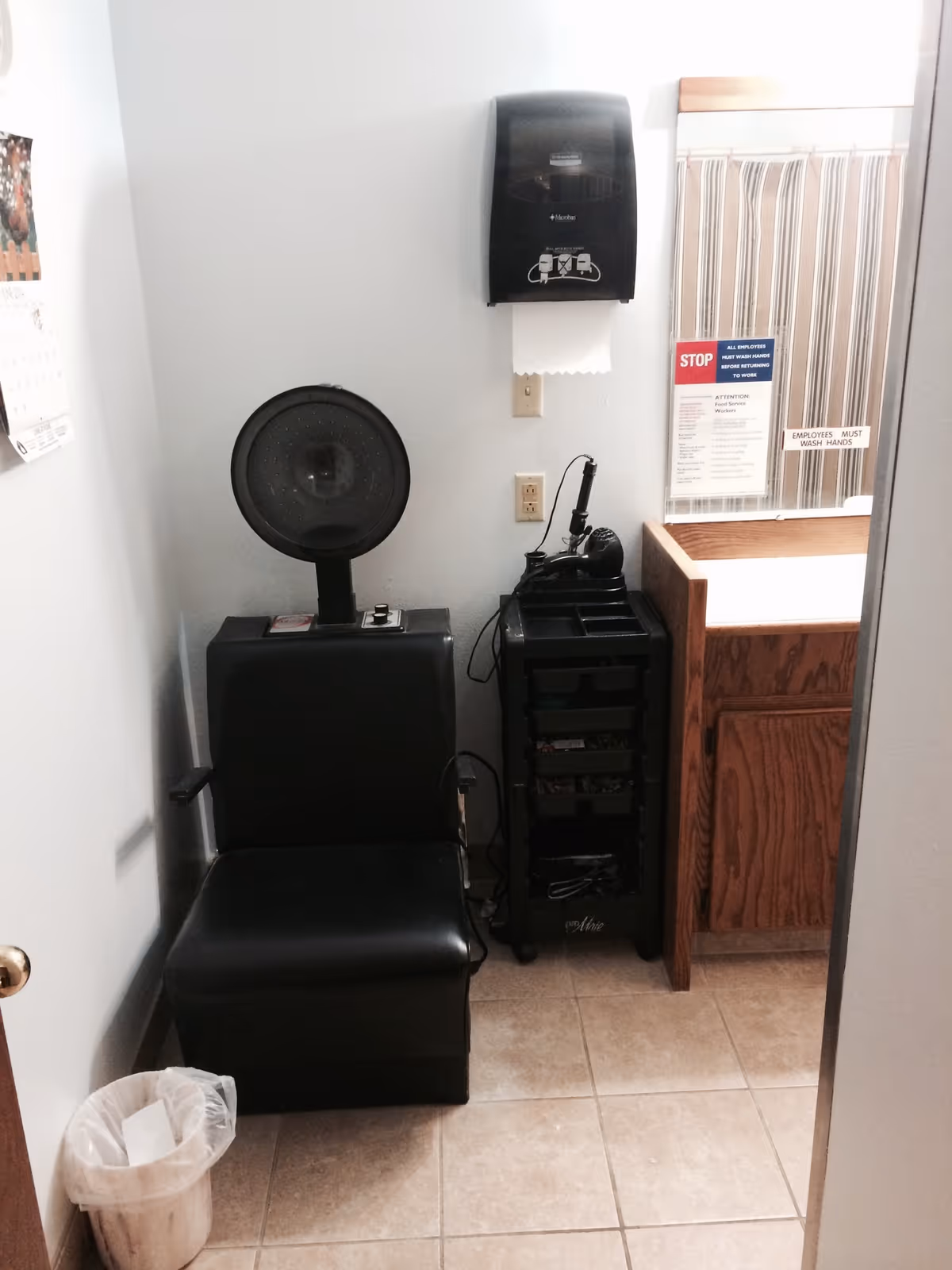 Small interior room with a black salon chair under a hooded hair dryer next to a rolling tool cart and a sink/vanity.