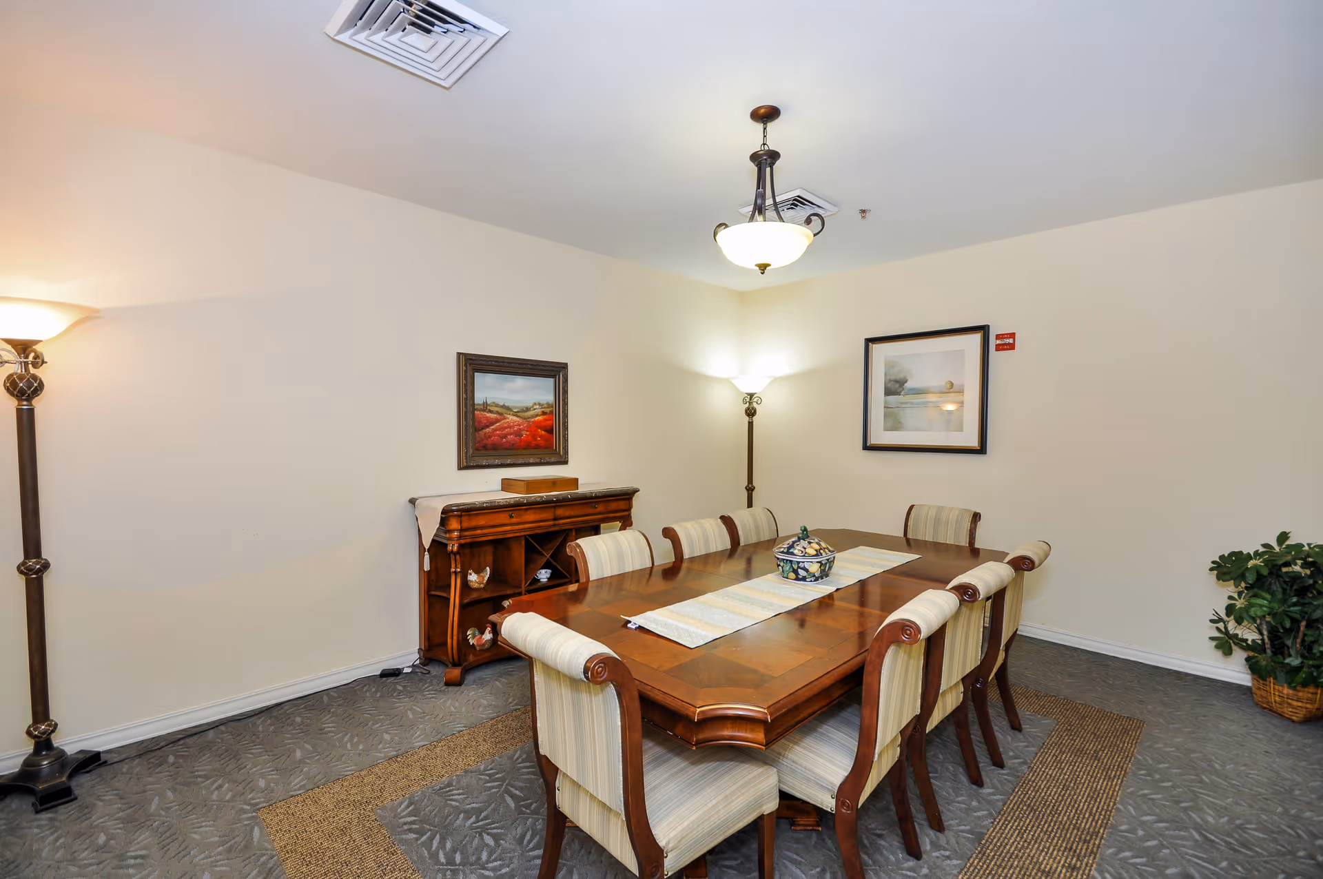 A dining room with a long wooden table surrounded by eight upholstered chairs. The table has a decorative runner and a centerpiece bowl. The room features two floor lamps, two framed paintings on the walls, a wooden sideboard with decorative items, and a potted plant in the corner. The walls are light-colored and the carpet has a patterned design.