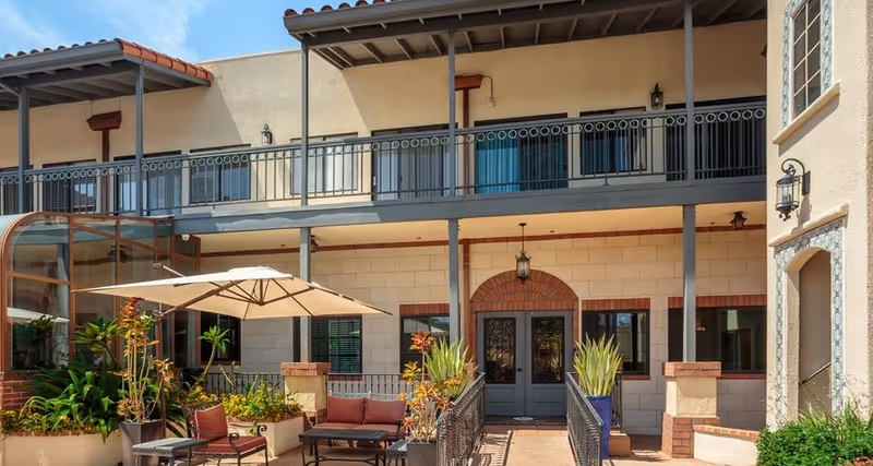 Sunny courtyard with patio seating, umbrella, planters and a two-story stucco building facade with a balcony.