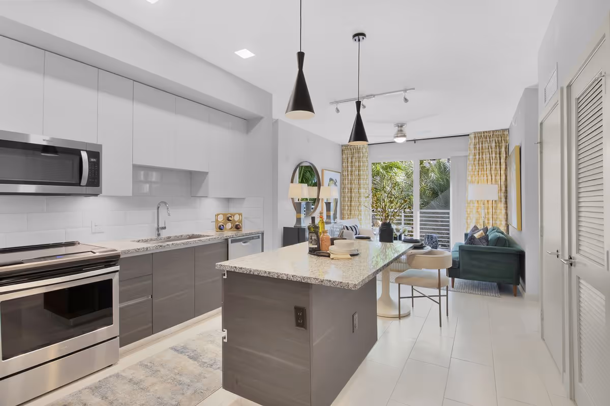Modern kitchen and living room area with white and gray cabinetry, stainless steel appliances, a granite countertop island with bar stools, pendant lights, and a green sofa near large windows with yellow patterned curtains overlooking greenery outside.