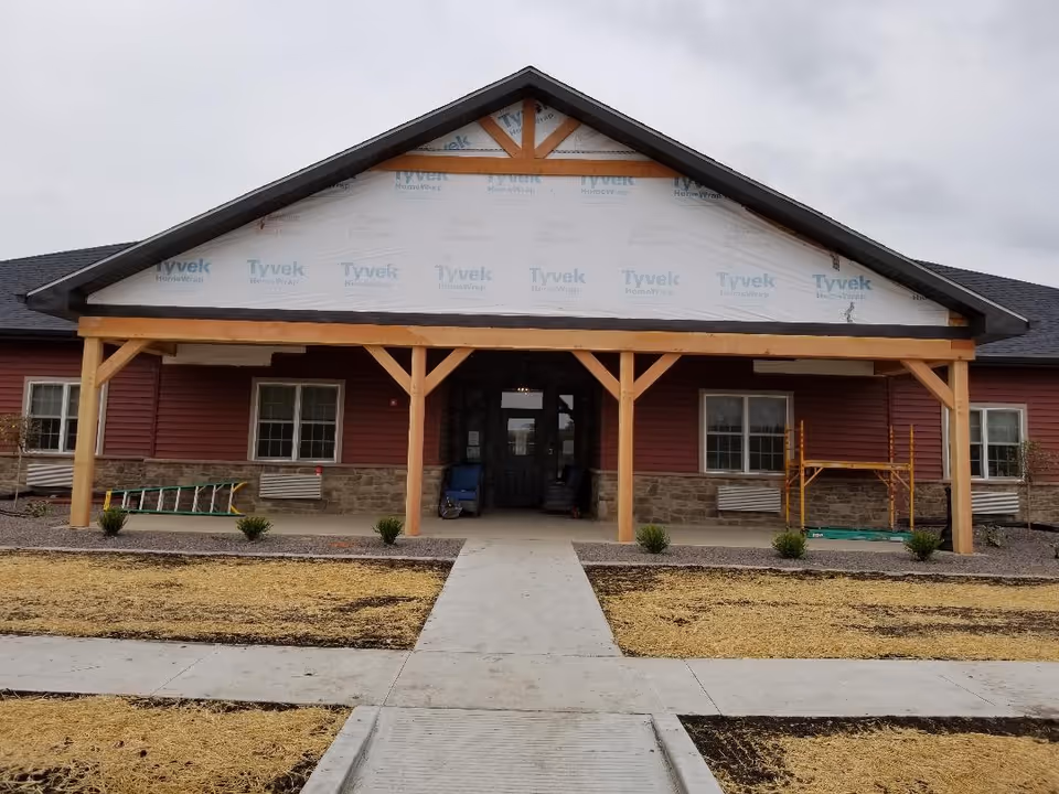 Front entrance of a single-story building with a covered wooden porch under construction and Tyvek housewrap visible on the gable.