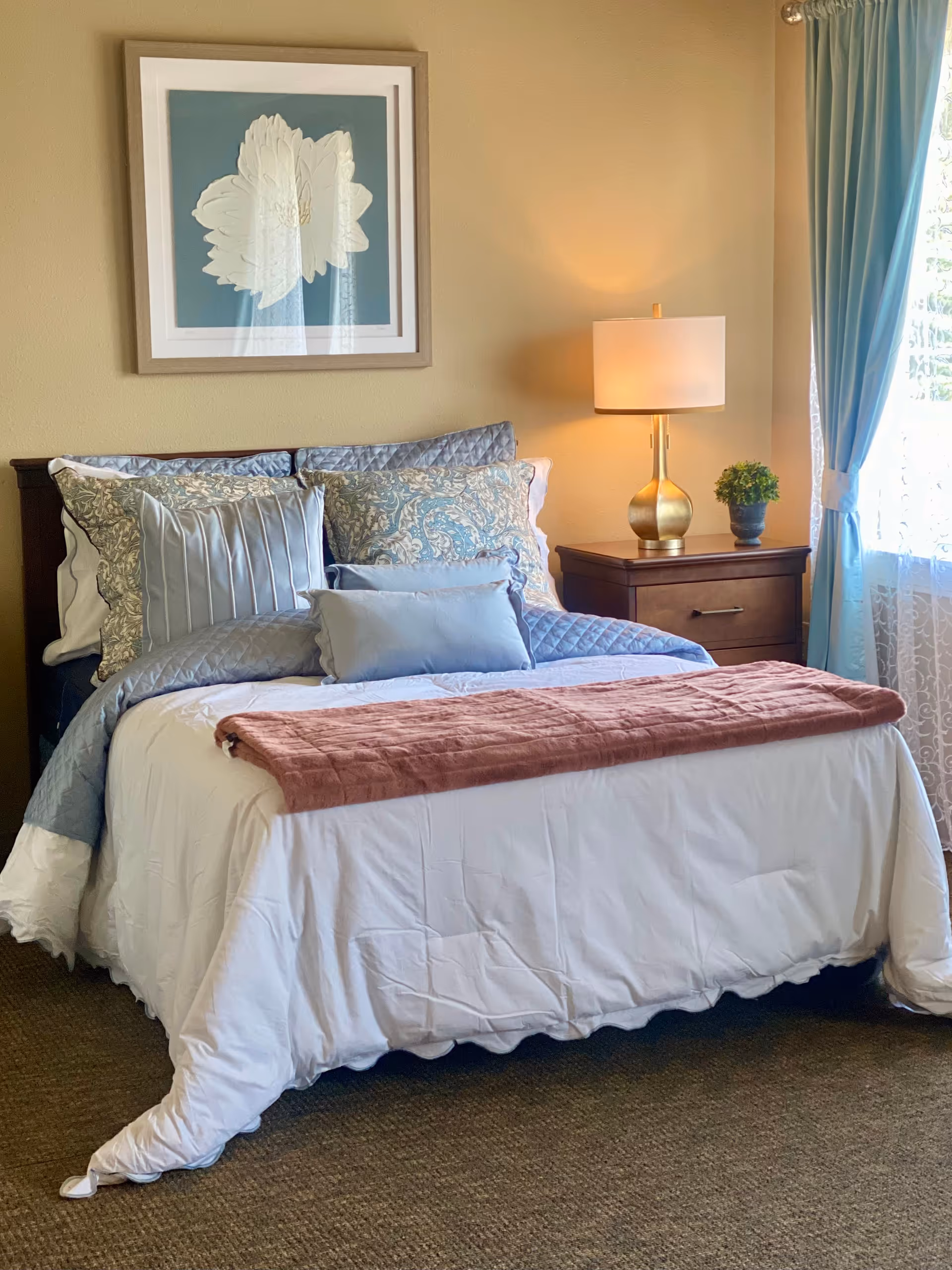A neatly made bed with multiple pillows and a white comforter in a bedroom. There is a wooden nightstand with a gold lamp and a small potted plant next to the bed. A framed floral artwork hangs on the wall above the bed. Light blue curtains frame a window with sheer white curtains allowing natural light into the room.
