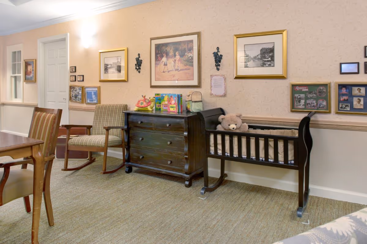 Cozy common room with chairs, a wooden dresser, a small crib holding a teddy bear, and framed pictures on the wall.