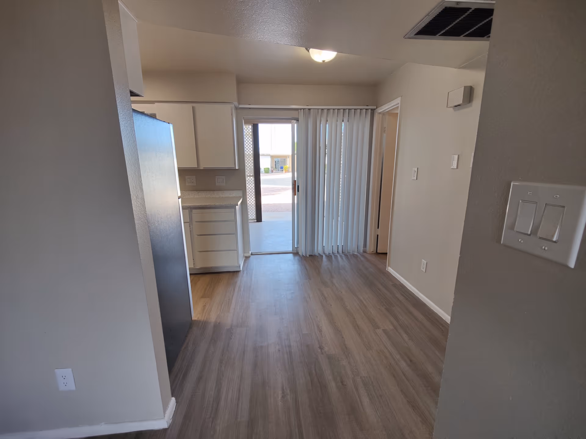 Interior view of an apartment with wood flooring, white walls, and a small kitchen area on the left with white cabinets and a refrigerator. There is a sliding glass door with vertical blinds leading to an outdoor area.