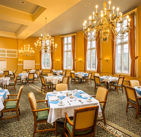 A spacious dining room with multiple tables covered in white tablecloths and set with cups, saucers, and silverware. The room features large windows with gold curtains, ornate gold chandeliers hanging from the ceiling, and patterned carpet flooring. Wooden chairs with green cushions surround each table.