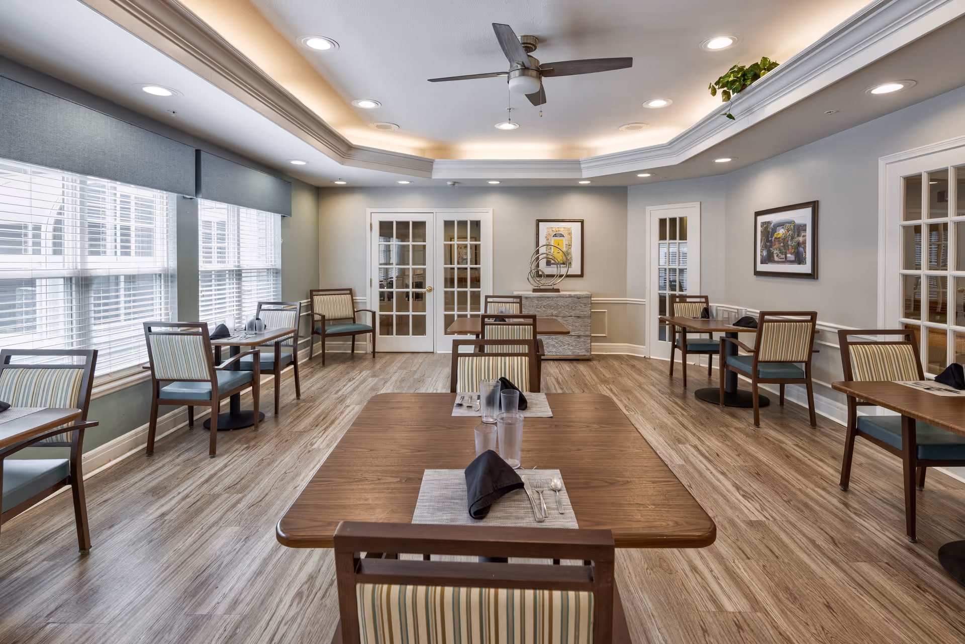 A bright dining room with several wooden tables and chairs arranged neatly. Each table is set with placemats, black napkins, utensils, and glasses. Large windows with blinds allow natural light to fill the room. The ceiling features recessed lighting and a ceiling fan. The walls are decorated with framed artwork and a decorative piece on a sideboard.