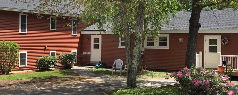 Exterior view of a red building with white trim at Assisted Living at Pine Hill Retirement Home. The building is surrounded by trees, bushes, and flowering plants. There are two white doors visible, a paved walkway, and a white plastic chair near the center.