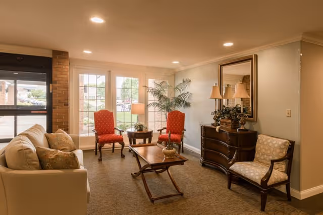 A cozy lobby seating area with a sofa, upholstered armchairs, a coffee table, sideboard with lamps and a large window letting in daylight.