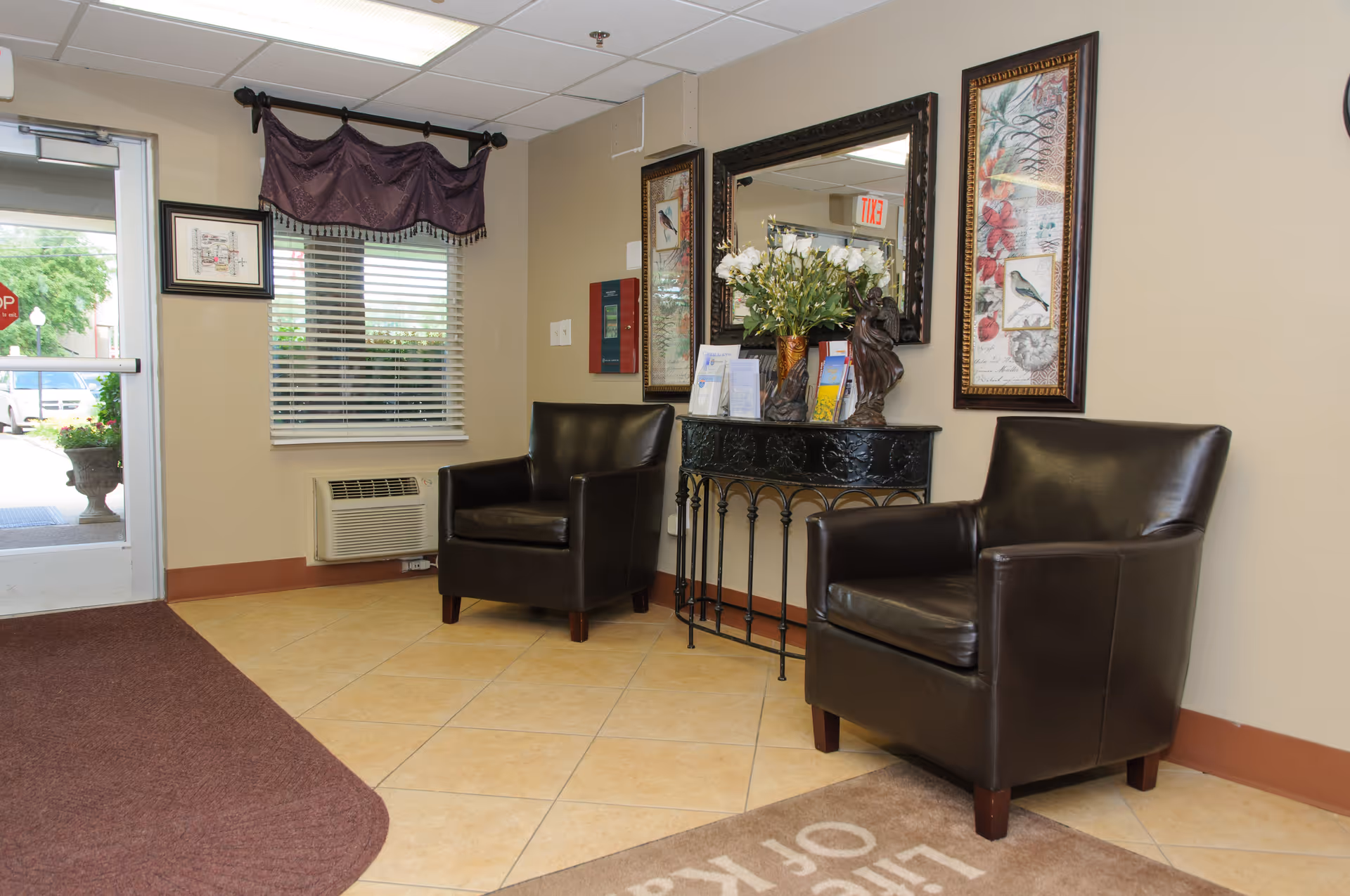 Entrance area of a facility with two dark brown leather armchairs, a decorative black console table with a vase of white flowers and brochures, a large mirror, framed artwork on the walls, a window with blinds and a purple valance, and a glass door leading outside.