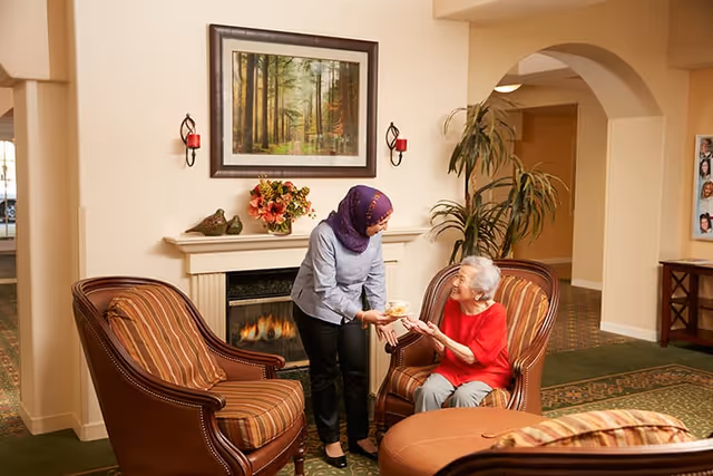 A woman wearing a purple headscarf and blue shirt is handing a cup to an elderly woman in a red top seated in a striped armchair in a cozy living room area with a fireplace, framed forest painting, and indoor plants.