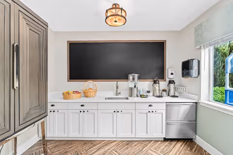 A clean and modern kitchenette area with white cabinets, a stainless steel sink, and a countertop holding a water dispenser, coffee pots, and baskets. Above the countertop is a large empty blackboard framed in wood. To the right, there is a window with a view of greenery outside. The floor has a wood-patterned design, and a light fixture hangs from the ceiling.