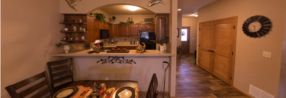 Interior view of a retirement community apartment showing a dining table set with plates and placemats in the foreground, a kitchen with wooden cabinets, a black refrigerator, and various decorative items in the background, and a hallway leading to a door with wooden double closet doors and a wall clock on the right.