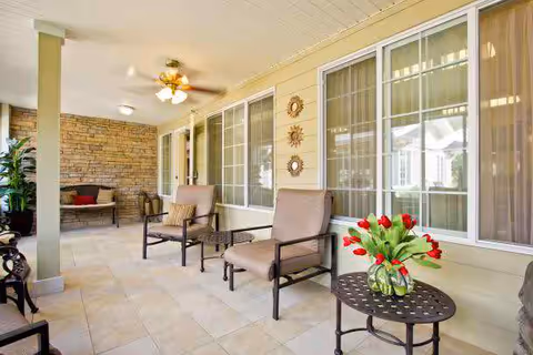 Covered patio with cushioned chairs, side tables and a vase of red tulips outside large windows.