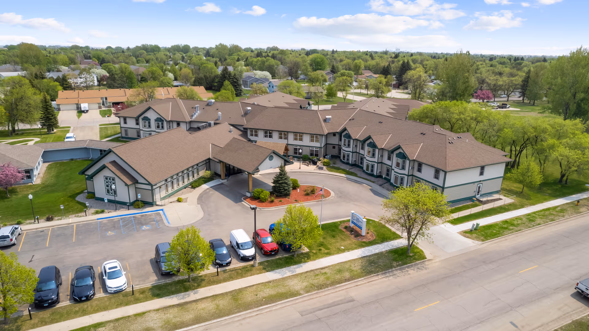 Aerial view of Good Samaritan Society - Heritage Grove, a large senior living facility with multiple connected buildings featuring brown roofs and light-colored walls with green trim. The facility is surrounded by trees and greenery, with a parking lot in front containing several cars. The sky is partly cloudy with blue patches.