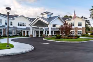 Front exterior view of Orchard Estate of Woodbury, a two-story building with a covered entrance, stone and siding facade, a flagpole with an American flag, and a curved driveway surrounded by landscaped greenery.