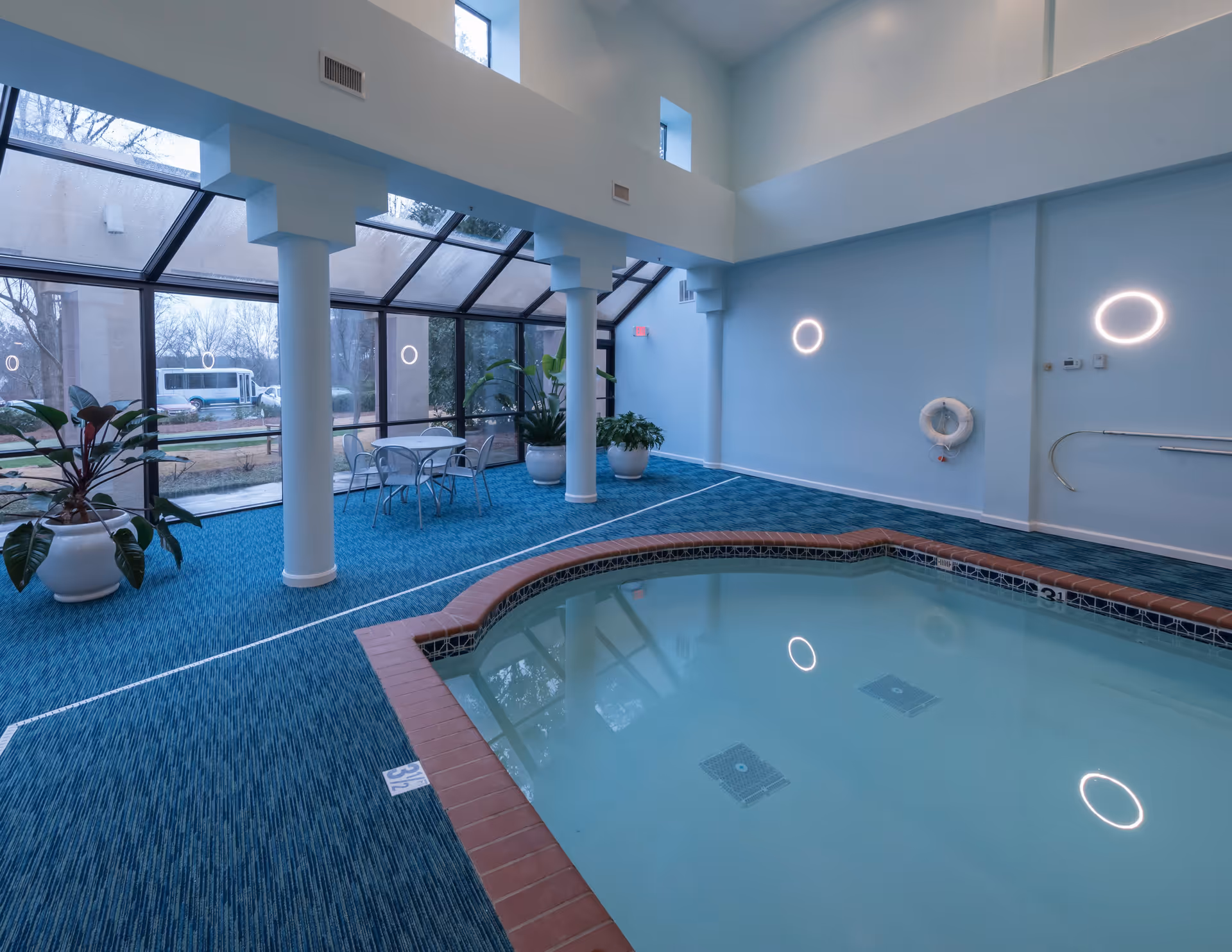 Indoor swimming pool area with blue carpeted floor and large windows letting in natural light. There are white columns, potted plants, a small table with chairs, and circular wall lights. The pool has a brick edge and a life preserver mounted on the wall.