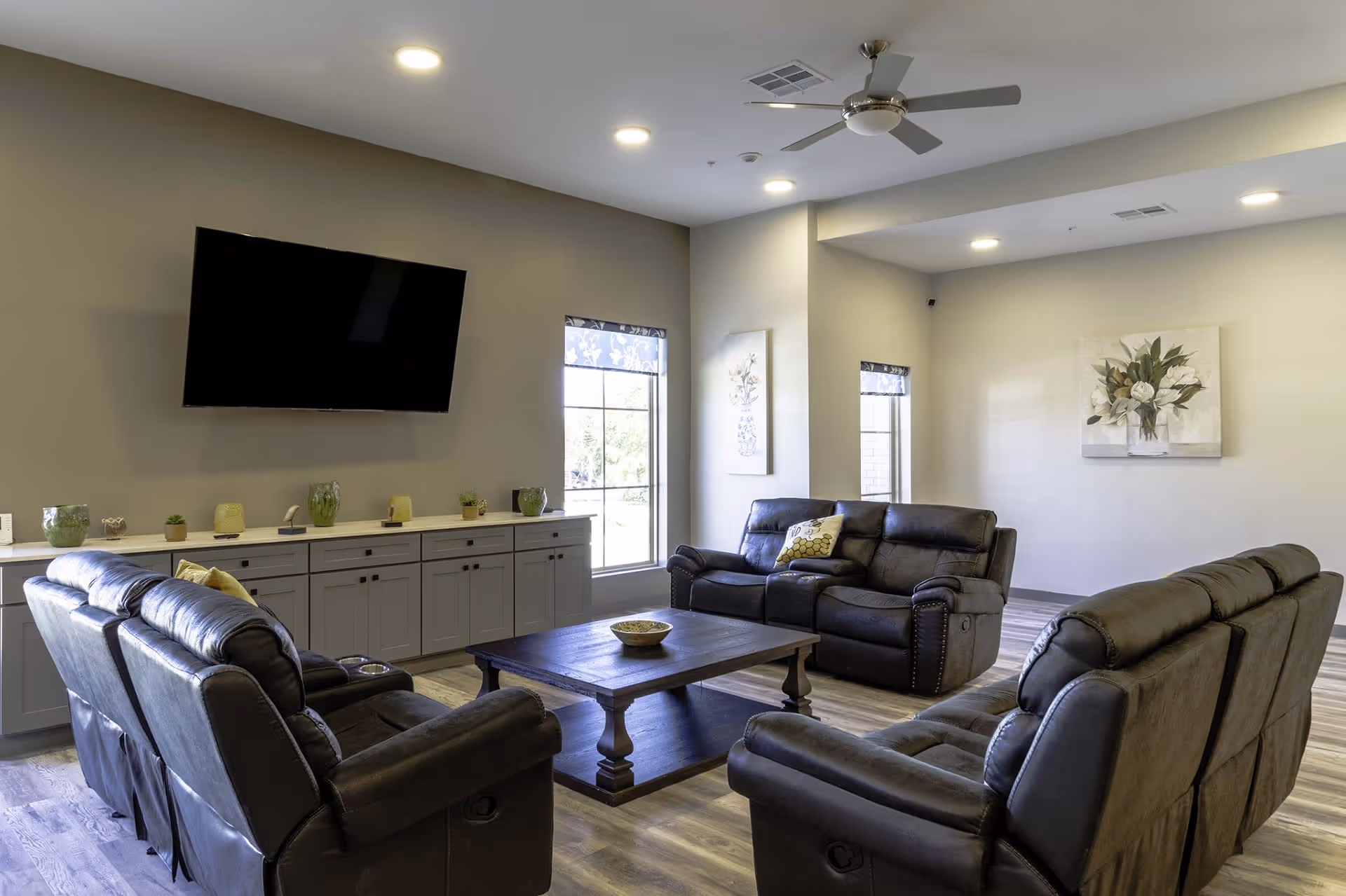 A cozy living room with three dark leather reclining sofas arranged around a wooden coffee table. A flat-screen TV is mounted on the wall above a long cabinet decorated with small plants and decorative items. Two large windows with floral-patterned blinds allow natural light to fill the room. The walls are adorned with floral artwork, and recessed ceiling lights and a ceiling fan provide additional lighting and ventilation.