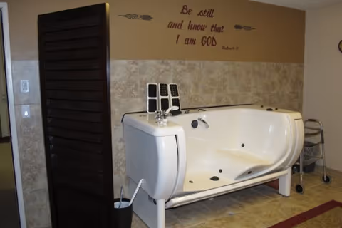 A walk-in bathtub in a tiled room with a privacy screen, a small rolling walker nearby, and a wall decal that reads 'Be still and know that I am GOD'.