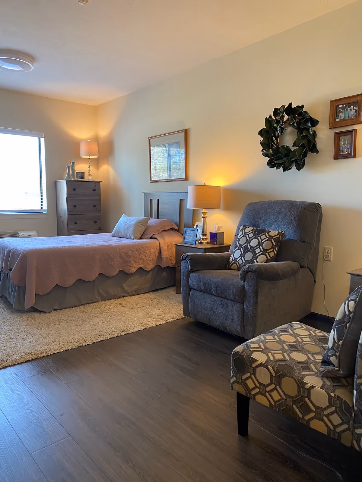 A cozy bedroom in The Meadows facility featuring a single bed with a pink quilt, a wooden dresser with a lamp and decorative items, a nightstand with a lamp and tissue box, a comfortable gray armchair with a patterned pillow, and a patterned accent chair. The room has light-colored walls adorned with a wreath and framed photos, a window with blinds, and wood flooring with a beige rug near the bed.