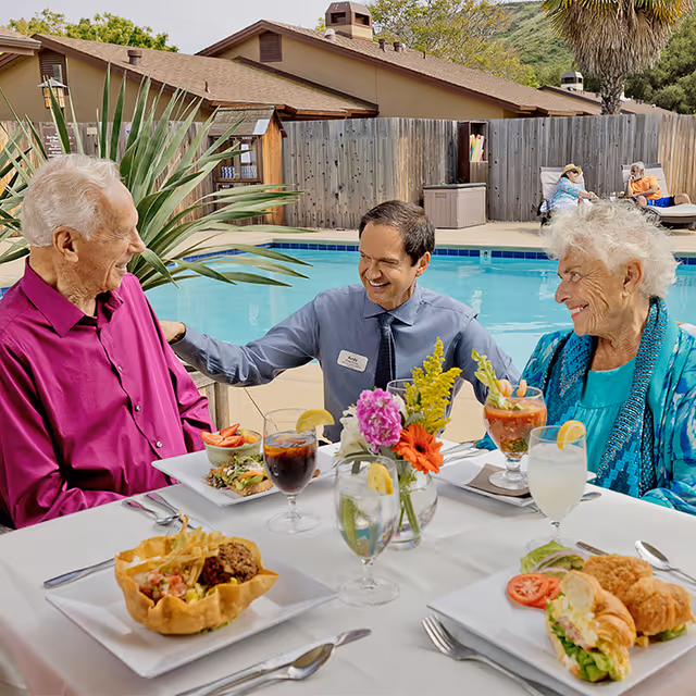 Two elderly residents and a staff member sit at a table beside a swimming pool, enjoying plated meals and drinks.
