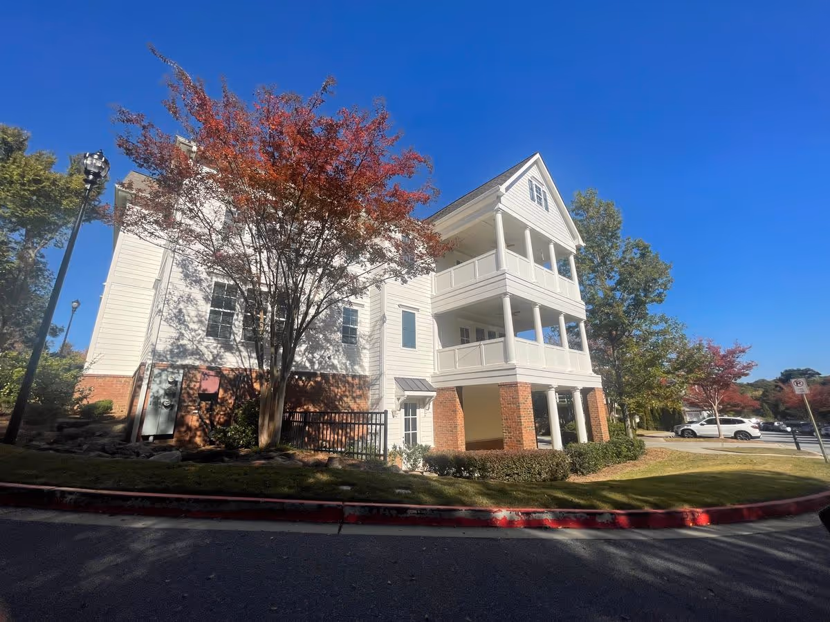 White three-story building with covered balconies and columns, trees in front and a parking area under a clear blue sky.