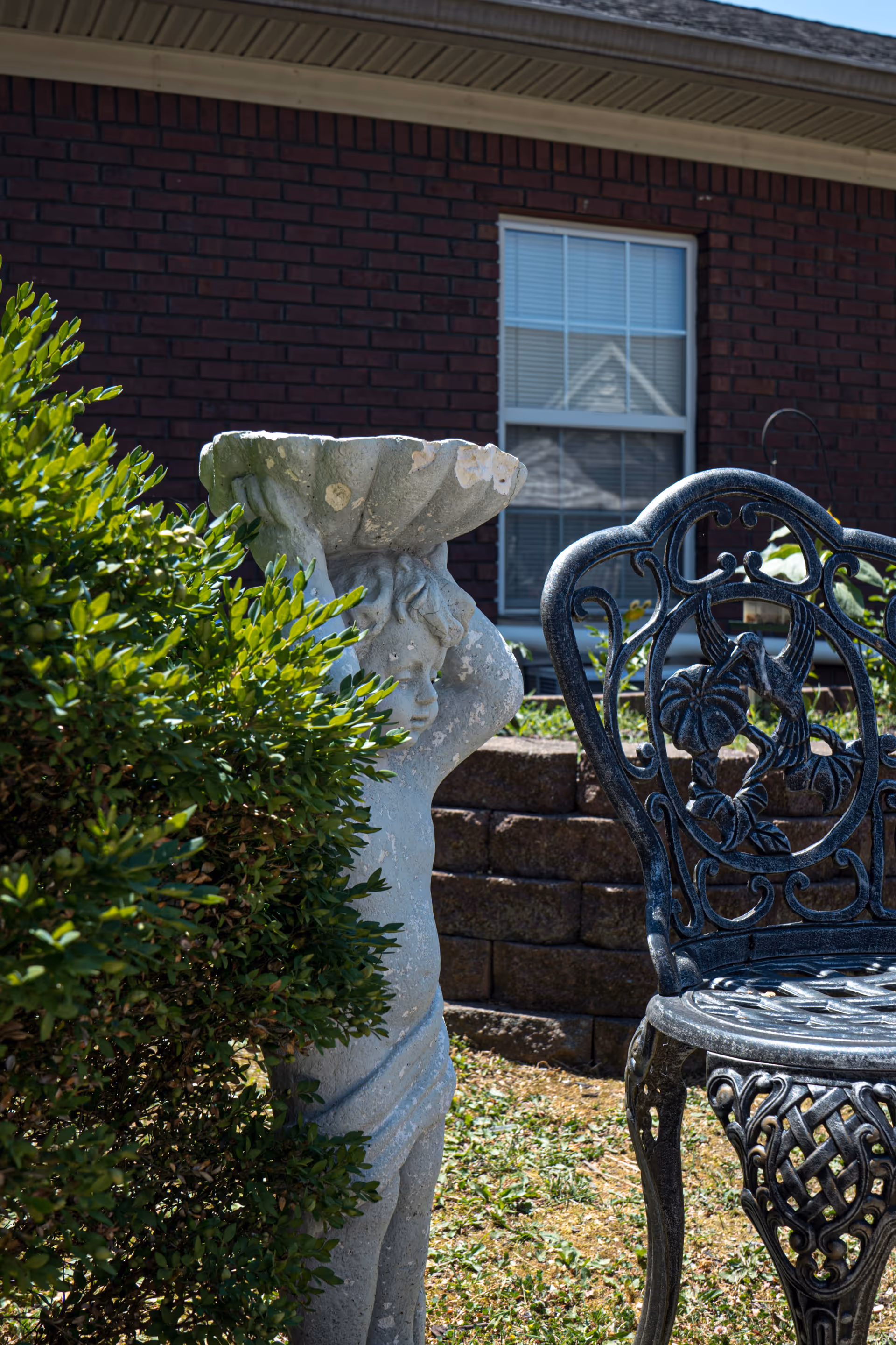 Outdoor garden area featuring a stone statue of a cherub holding a bowl, partially obscured by green bushes, with a decorative black metal chair nearby and a brick building with a window in the background.
