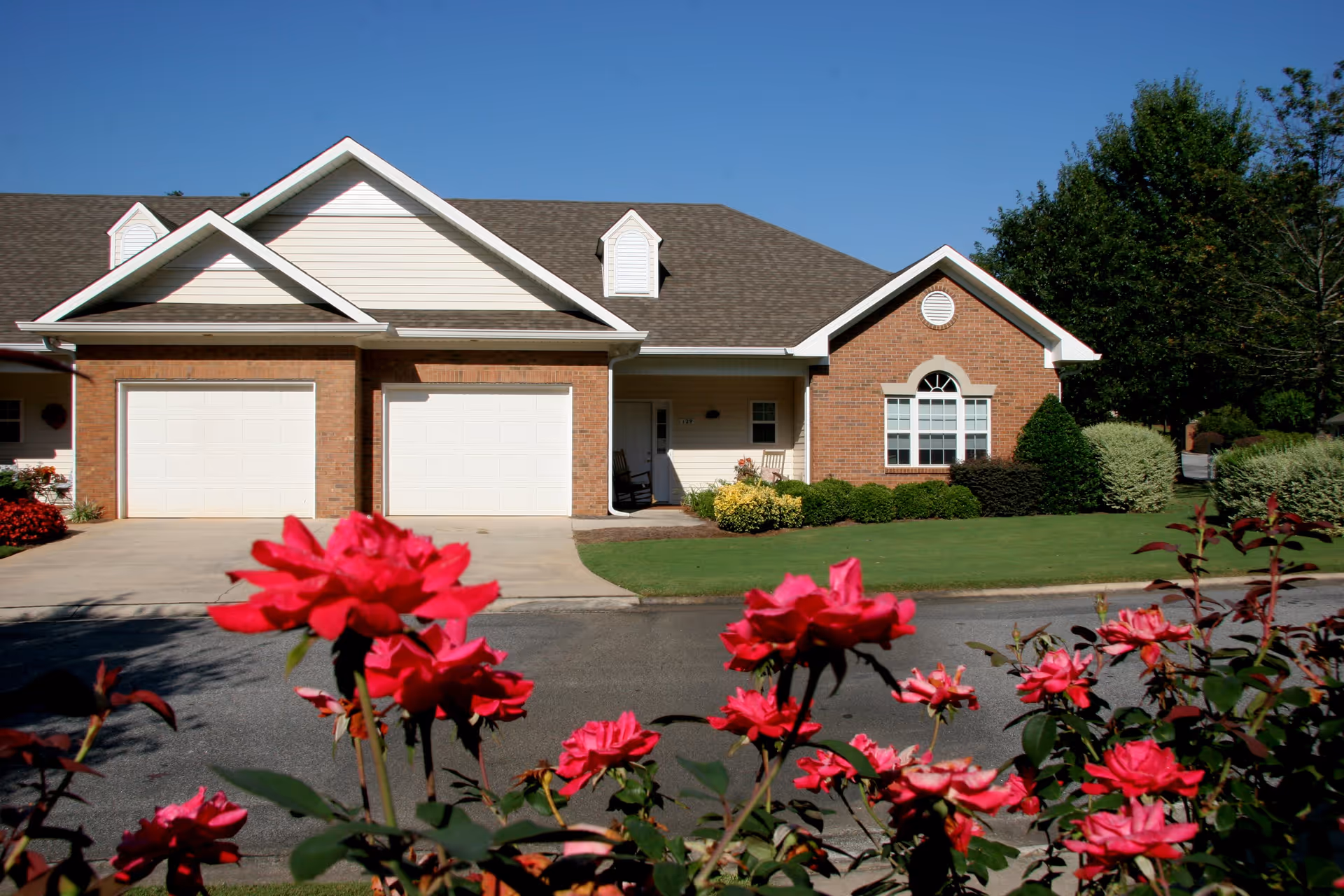 Exterior view of a single-story residential building with two white garage doors, a small front porch with a rocking chair, and a well-maintained lawn. Bright pink flowers are in the foreground, and trees are visible in the background under a clear blue sky.
