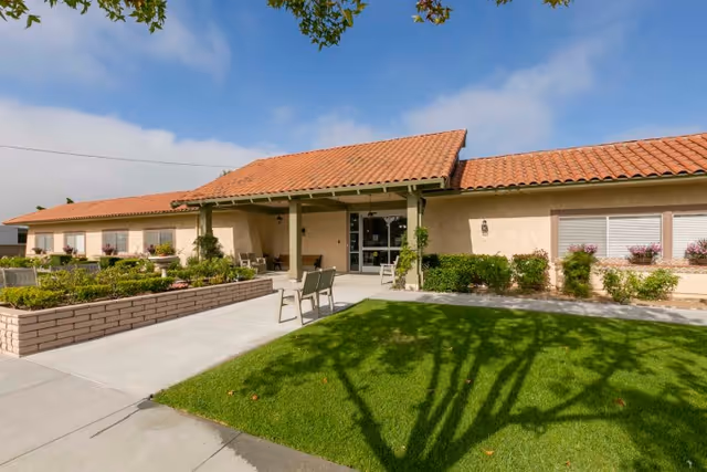 Exterior view of Camarillo Healthcare Center showing a single-story building with a red tile roof, beige walls, and a covered entrance. There is a well-maintained lawn and garden area with benches and potted plants near the entrance under a clear blue sky.