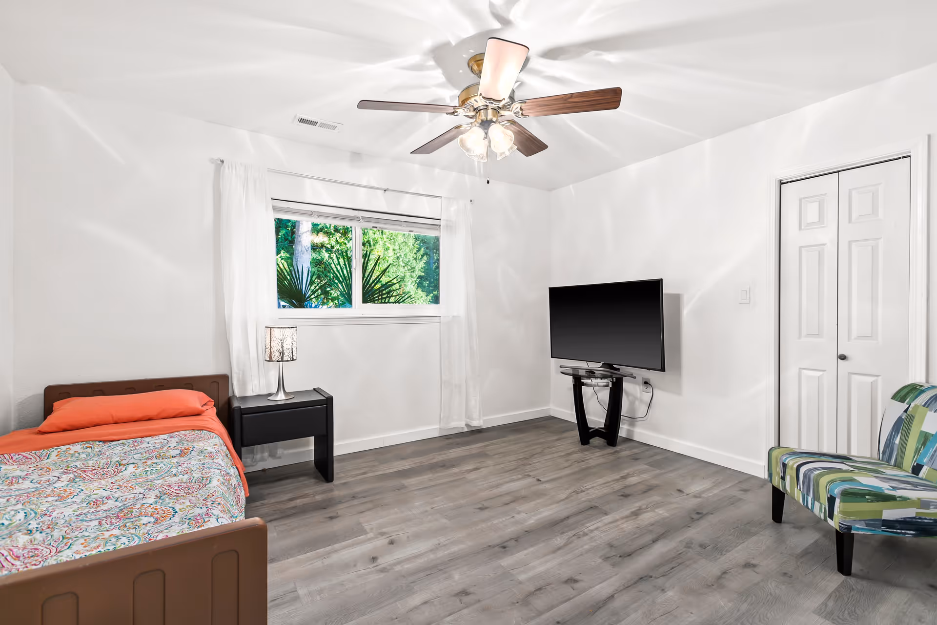 Bright, minimalist bedroom with a single bed, nightstand and lamp, wall-mounted TV, patterned accent chair, and a ceiling fan.