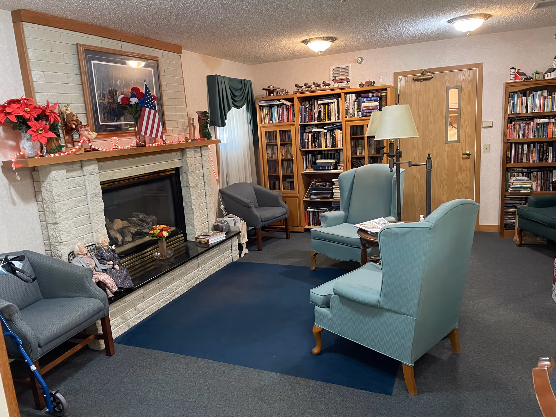 Cozy communal living room with a fireplace, bookshelves, and upholstered armchairs arranged around a central rug.