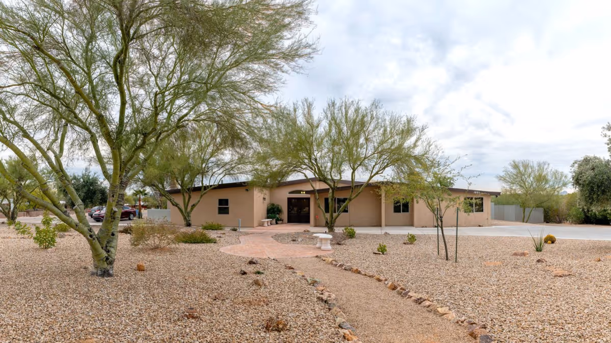 Single-story beige building with a flat roof surrounded by desert landscaping including gravel, small bushes, and several leafless trees under a cloudy sky. A curved brick pathway leads to the entrance with a bench and a small white table nearby. A few cars are parked to the left side of the building.