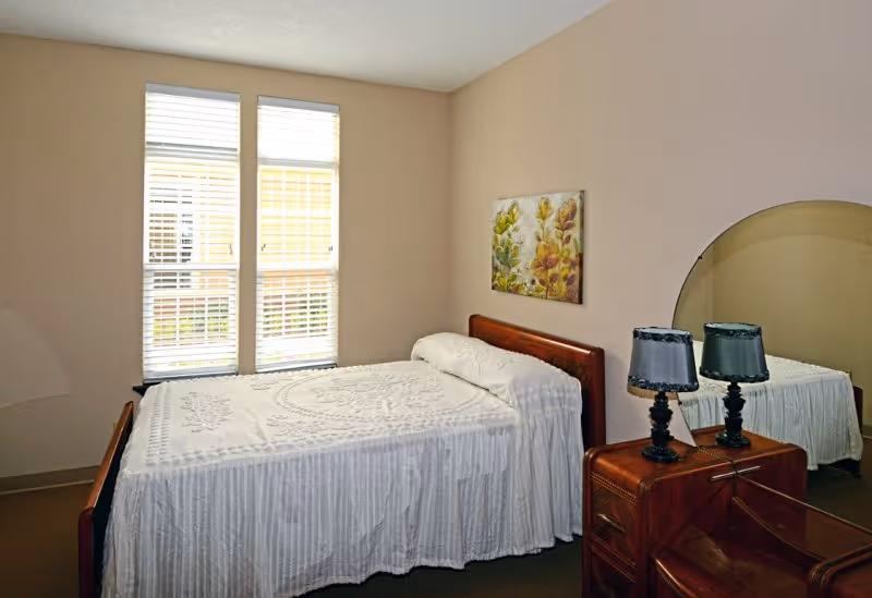 A simple bedroom with a bed covered in a white textured bedspread, positioned against a beige wall. There are two tall windows with white blinds letting in natural light. Next to the bed is a wooden nightstand with two black lamps and a large round mirror behind it. A floral painting hangs on the wall above the bed's headboard.
