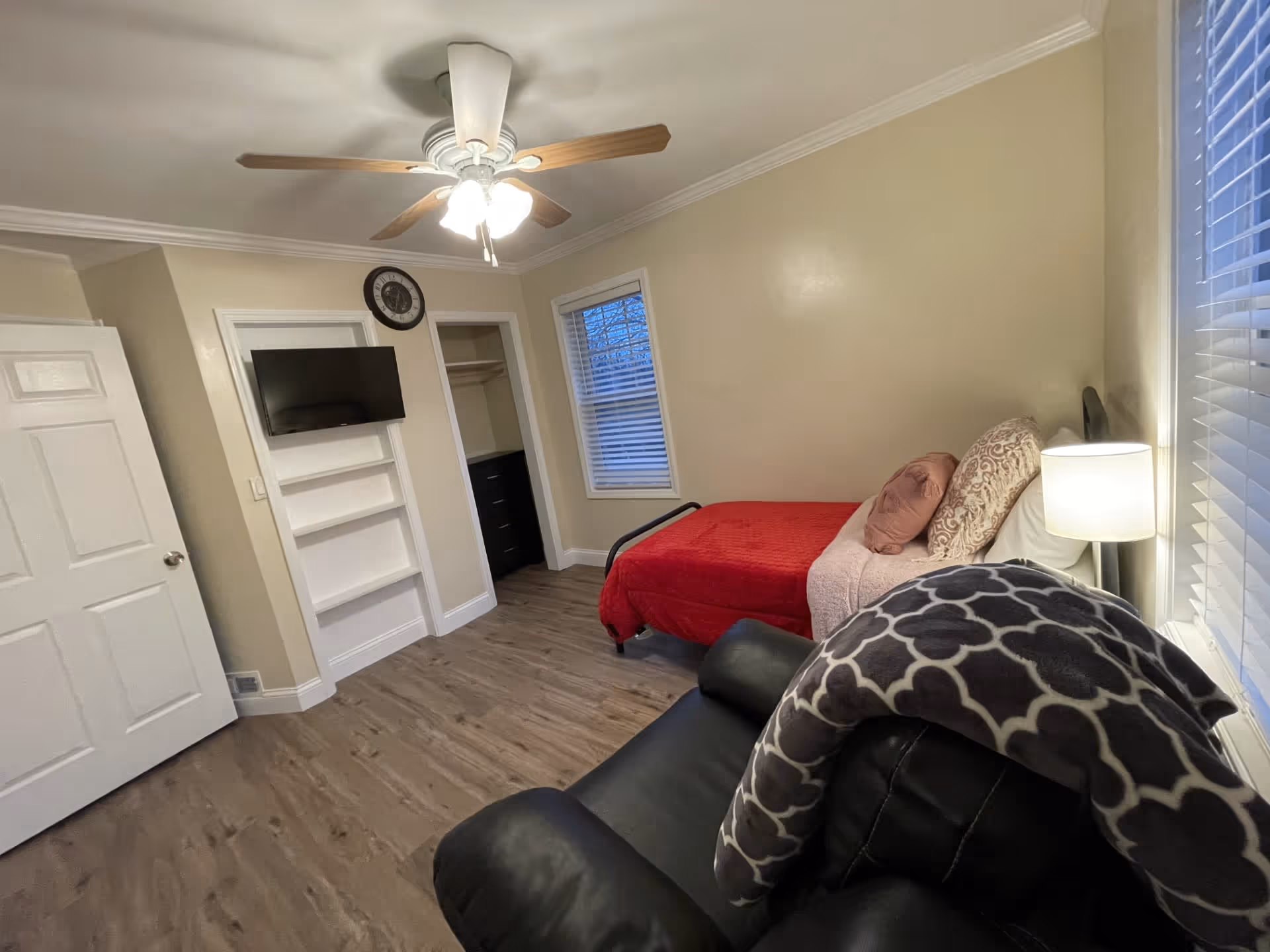 A small bedroom with a red-covered single bed along the right wall, a black recliner in the foreground, a wall-mounted TV, ceiling fan, and window blinds.