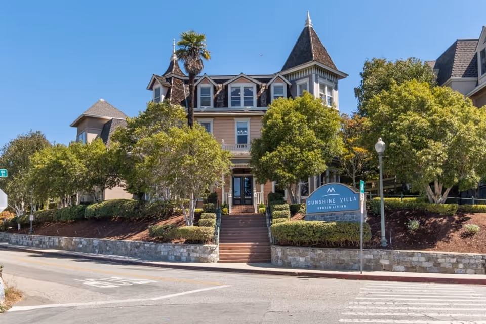 Exterior view of Sunshine Villa, a Merrill Gardens Community senior living facility, showing a large, elegant building with a steep roof and multiple windows, surrounded by well-maintained trees and landscaping under a clear blue sky.