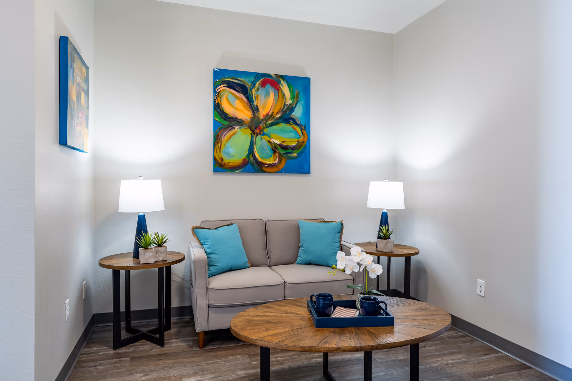 A cozy living room area with a beige loveseat adorned with two turquoise pillows, flanked by two round wooden side tables each holding a blue lamp and a small plant. A round wooden coffee table in front holds a blue tray with two blue mugs and a white orchid. The walls are white with two colorful abstract paintings, one large flower painting centered above the loveseat and a smaller one on the left wall. The floor is wood with a gray baseboard.