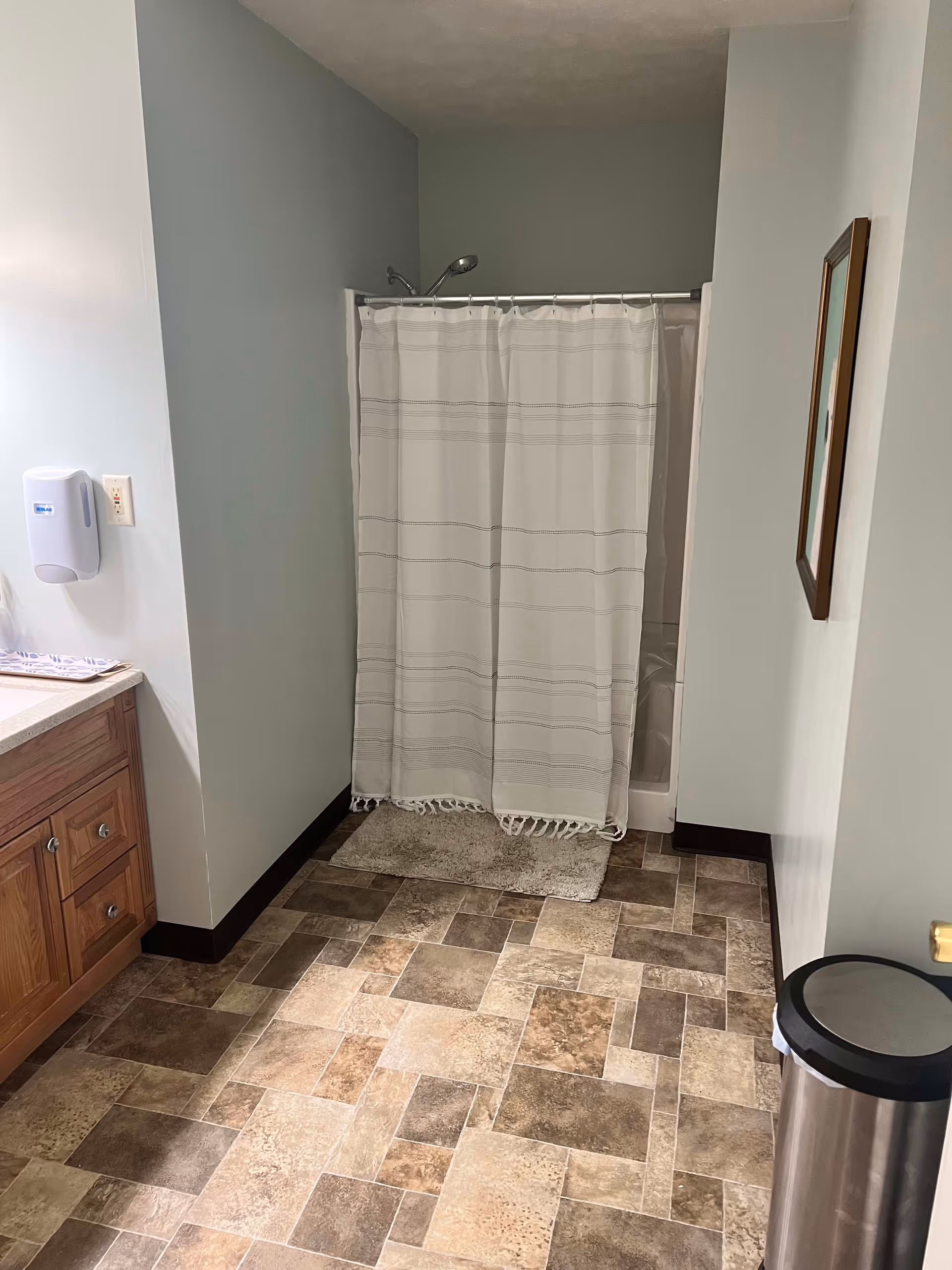 A bathroom with a shower area covered by a white curtain with horizontal stripes. The floor has a patterned tile design in shades of brown and beige. To the left, there is a wooden vanity with drawers and a soap dispenser mounted on the wall. On the right side, there is a trash can and a framed picture hanging on the wall.