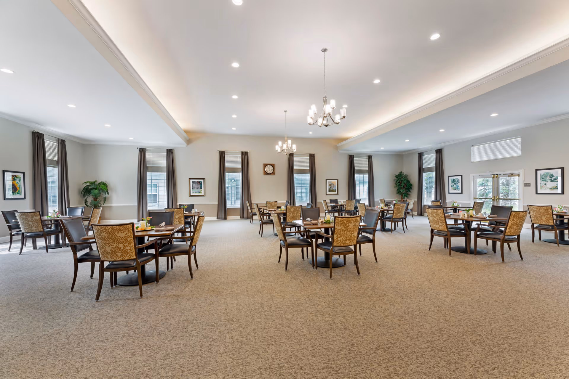 A spacious dining room in a senior living facility with multiple round tables and chairs arranged neatly. The room has large windows with dark curtains, light-colored walls, carpeted floors, and two chandeliers hanging from the ceiling. There are framed pictures on the walls and potted plants in the corners.