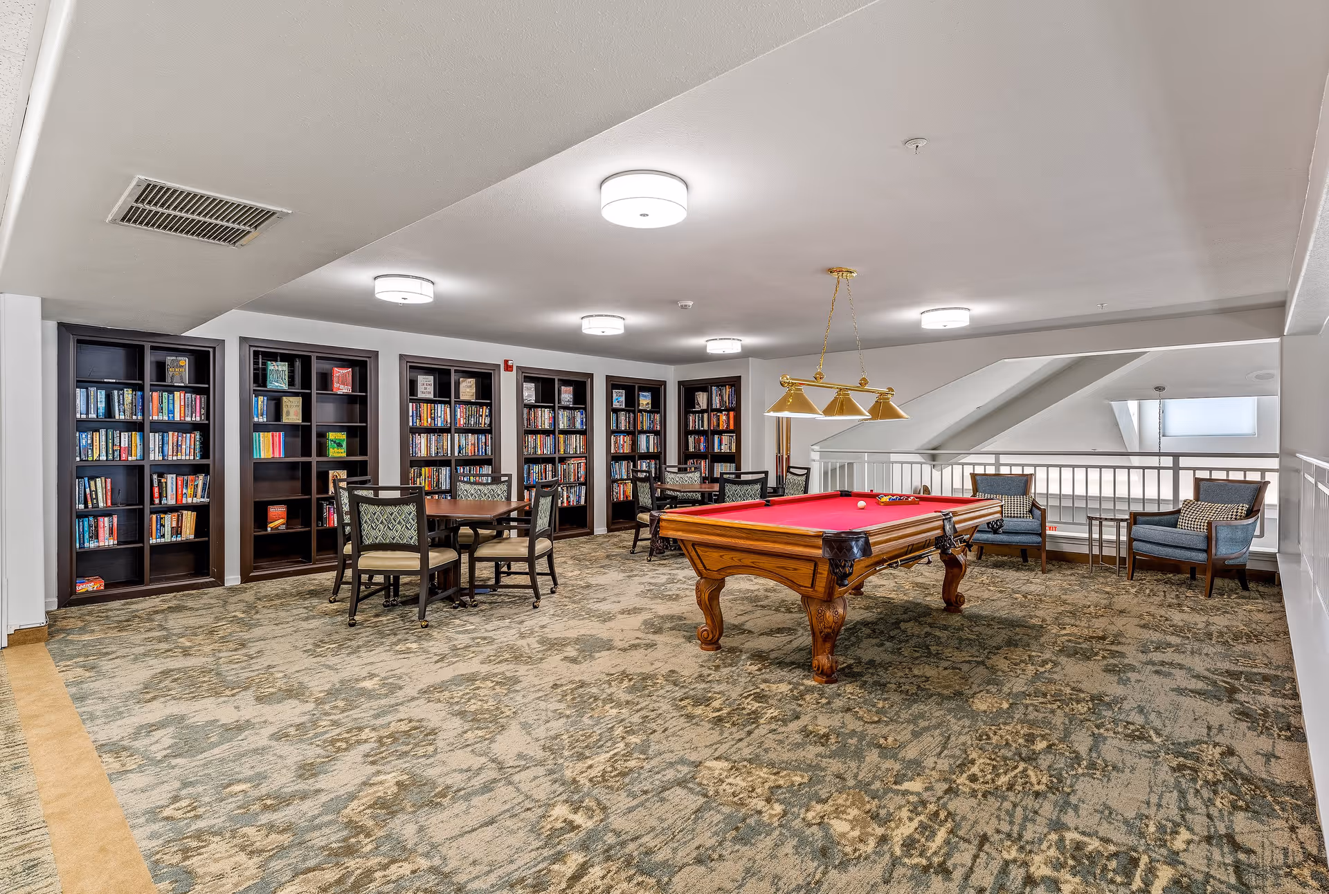 A spacious recreational room featuring a red felt pool table with a hanging light fixture above it, several bookshelves filled with books along the back wall, round tables with chairs, and a seating area with armchairs near a railing overlooking a lower level.