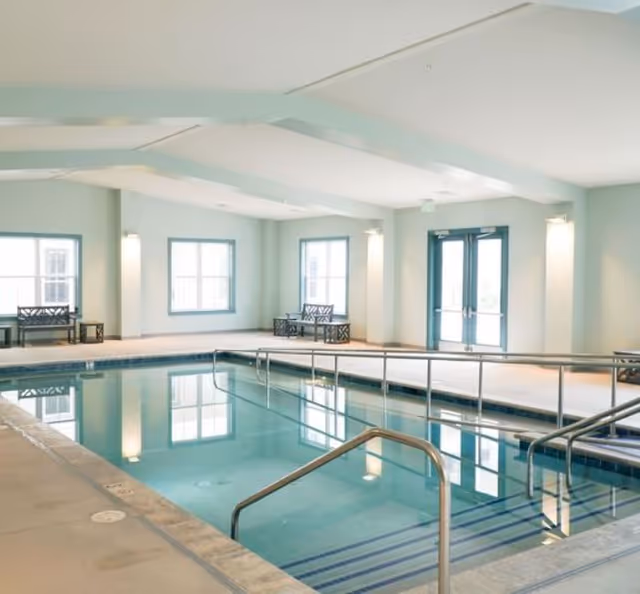 Bright indoor swimming pool with handrails, entry steps, benches and windows in a spacious room.