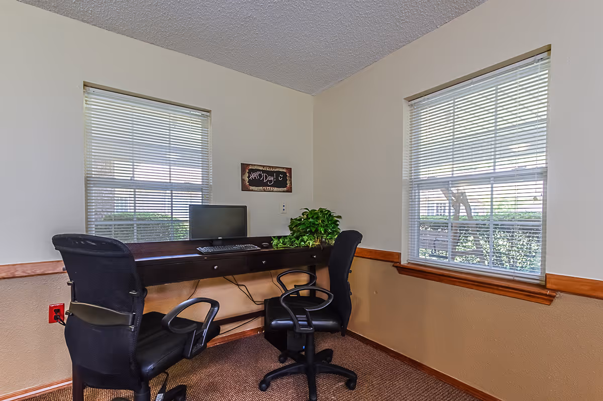 A small office area with two black office chairs and a dark wooden desk mounted to the wall. On the desk is a computer monitor, keyboard, and a green potted plant. Two windows with white blinds allow natural light into the room. A small wall decoration above the desk reads 'Happy Day!'. The walls are painted beige with a wooden chair rail and brown carpeted floor.