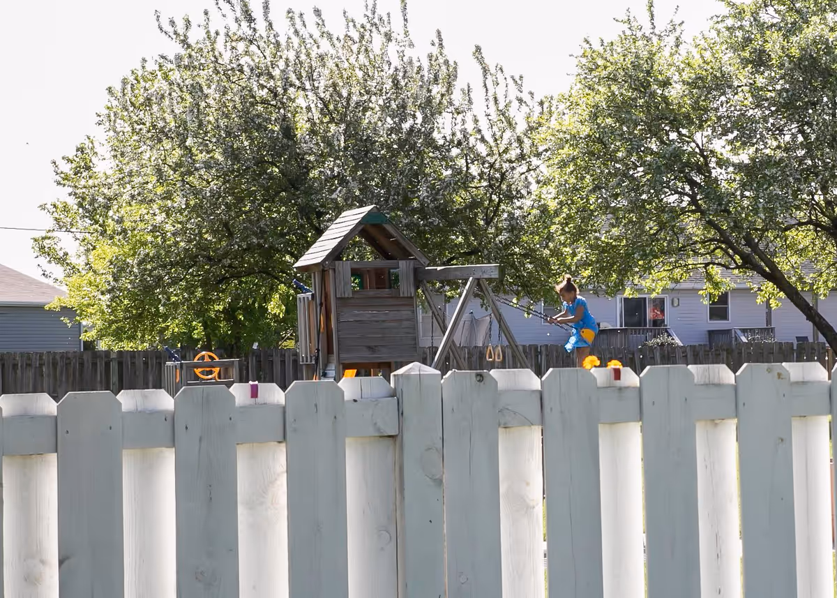 A child playing on a wooden playground set with swings in a backyard surrounded by a white wooden fence and green leafy trees.