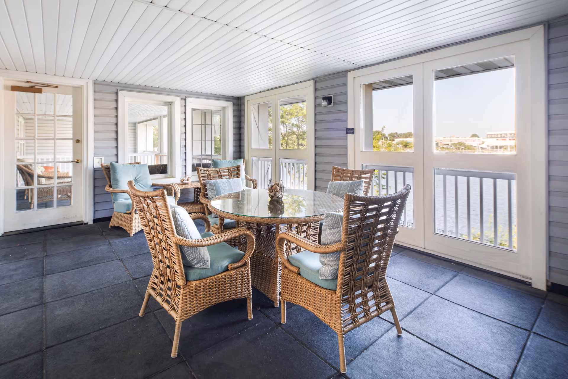 A bright enclosed patio area with a round glass-top wicker table surrounded by six wicker chairs with light blue cushions and pillows. The space has large windows and glass doors that offer a view of a body of water and greenery outside. The floor is tiled with large dark tiles, and the walls are covered with light gray siding.